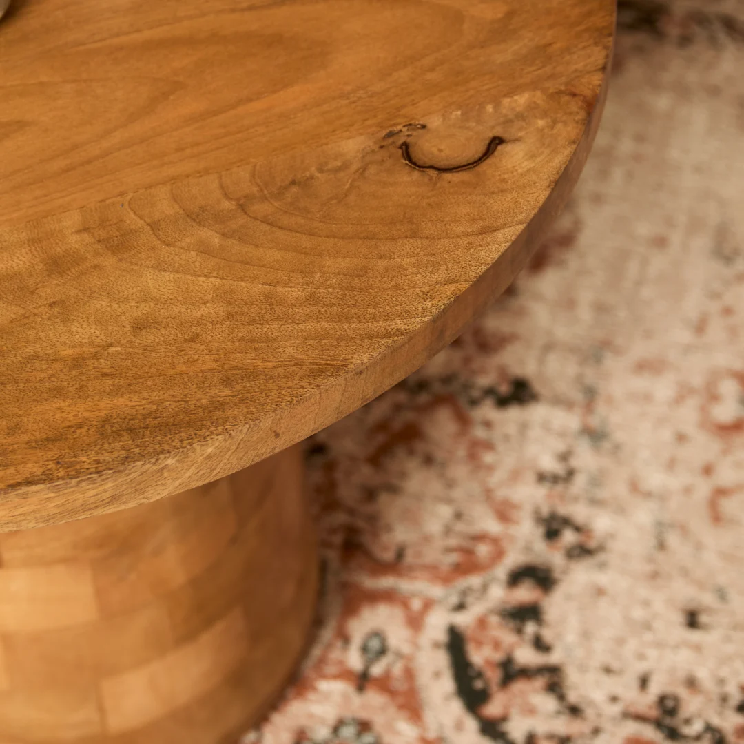 Een close-up van het ronde houten tafelblad van Timo, met zichtbare nerven en een kleine donkere knoest, boven een vloerkleed met rode, zwarte en crèmekleurige patronen.