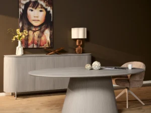 Ovale eettafel Solana in een kamer met houten vloer en donkere muren. Een dressoir, lamp en schilderij staan op de achtergrond. Zachte belichting.