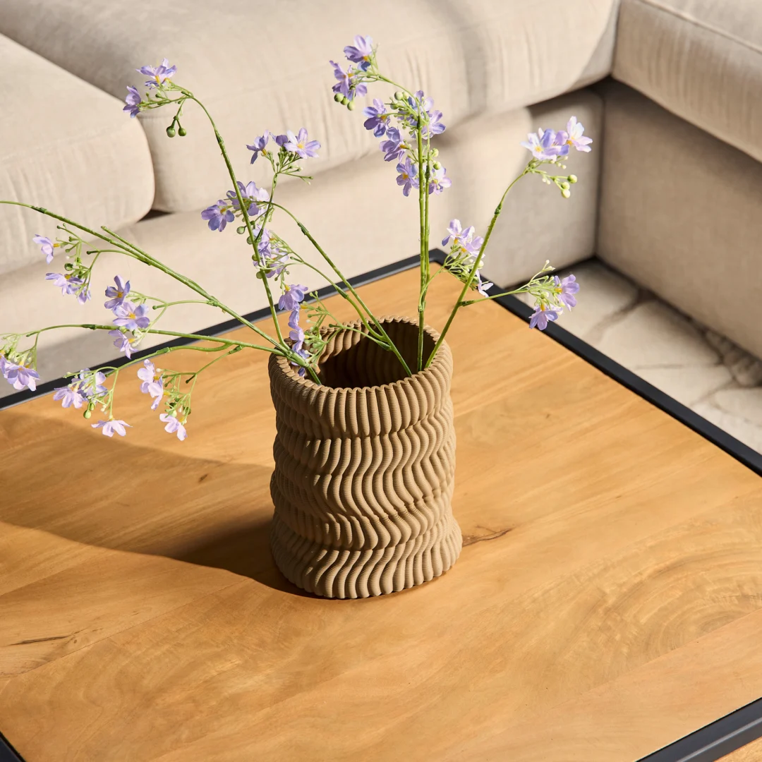 Een golvende, gestructureerde beige vaas met delicate paarse bloemen staat op de Salontafel 80 x 80 CM Rond Design, met een lichtgekleurde stoffen sectionele bank op de achtergrond.