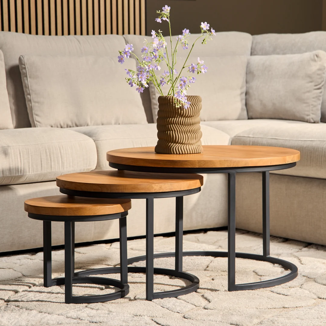 De Salontafel 70 x 45 CM Rond Nesting Set, met houten bladen en zwarte metalen frames, staat opgesteld voor een beige bank. Een gestructureerde vaas met paarse bloemen staat op de grootste tafel op een tapijt met crèmekleurig patroon.
