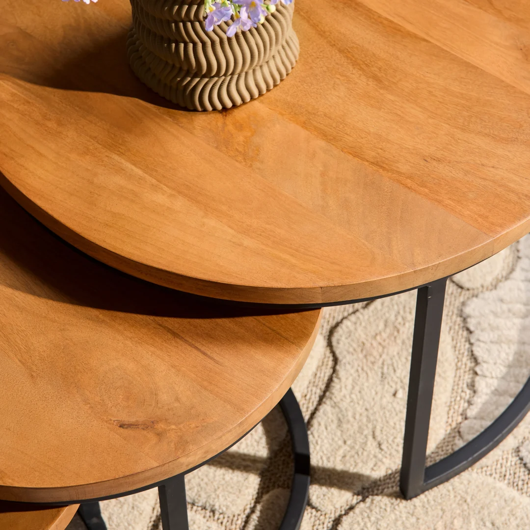 De Salontafel 70 x 45 CM Rond Nesting Set bestaat uit twee overlappende ronde houten tafels met zwarte metalen poten op een beige vloerkleed met textuur. Op de bovenste tafel staat een gegroefde vaas met paarse bloemen.