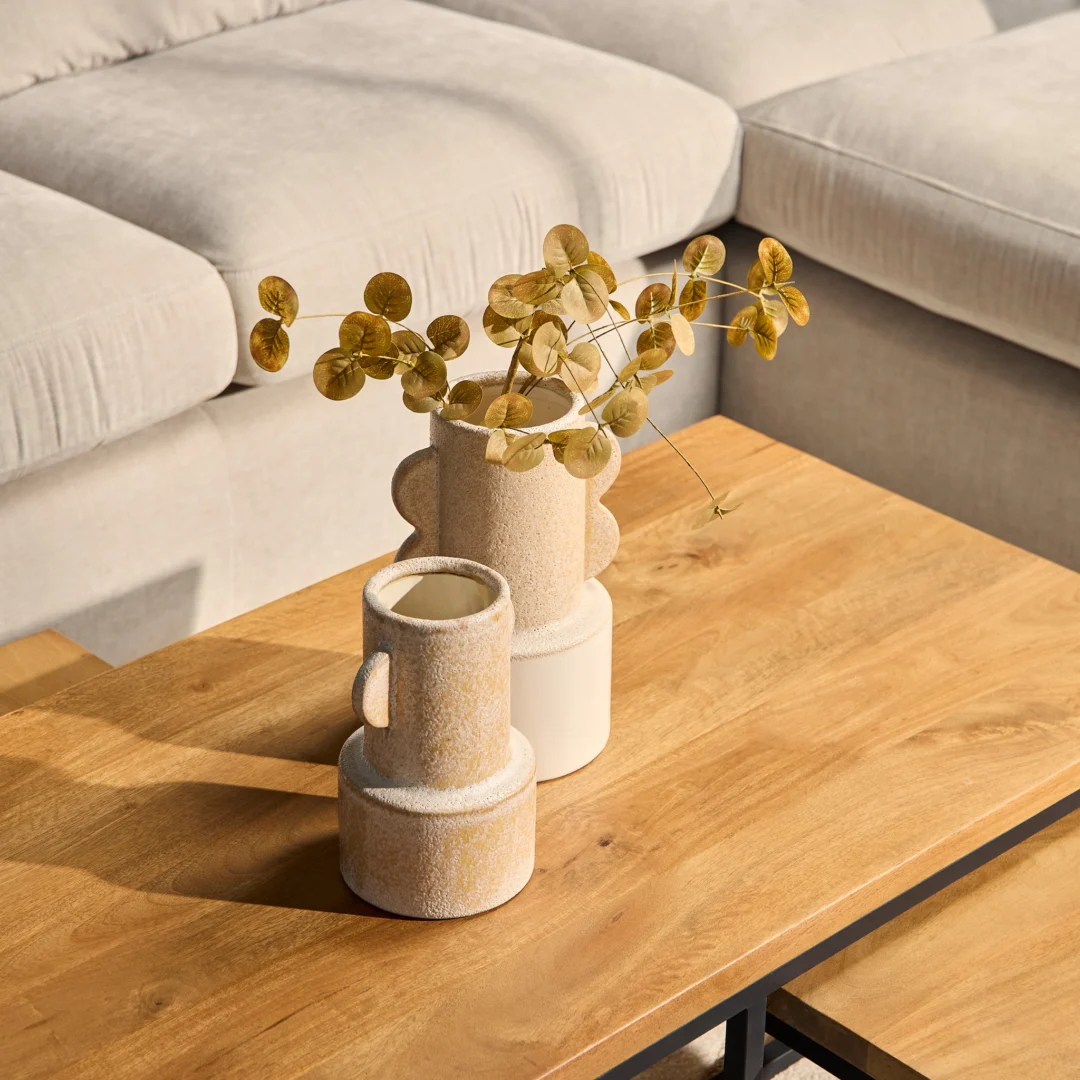 Twee beige keramische vazen met gedroogde ronde gouden bladeren staan op de Salontafel Roest 50 x 42 CM Rechthoek voor een lichtgekleurde bank. Zonlicht werpt zachte schaduwen en creëert een warme, minimalistische sfeer.