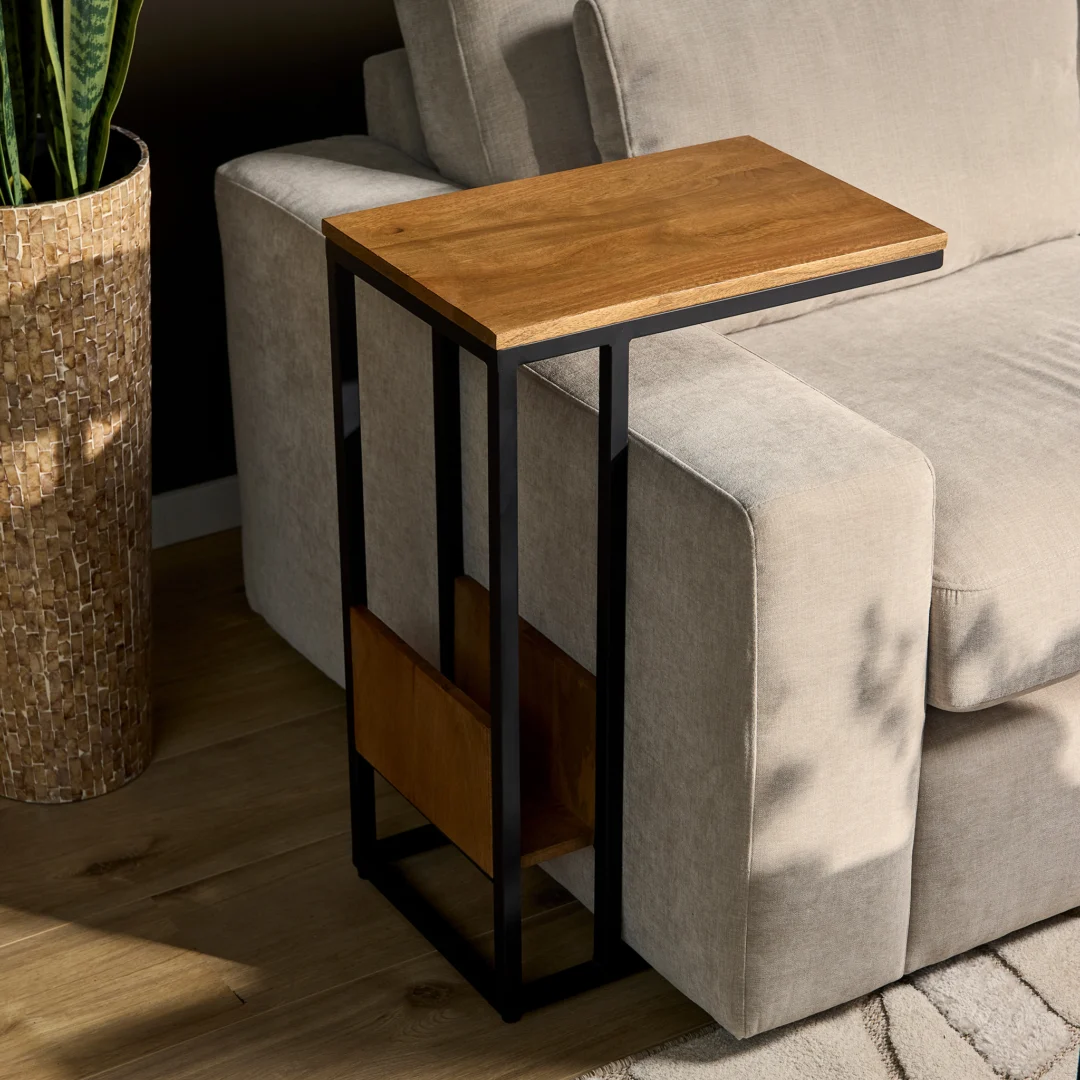 De Schuifregelaar Naturel Hoog, een moderne C-vormige bijzettafel met een houten blad en een zwart metalen frame, staat naast een lichtgrijze bank op een houten vloer, naast een hoge gevlochten mand met daarin een groene plant.