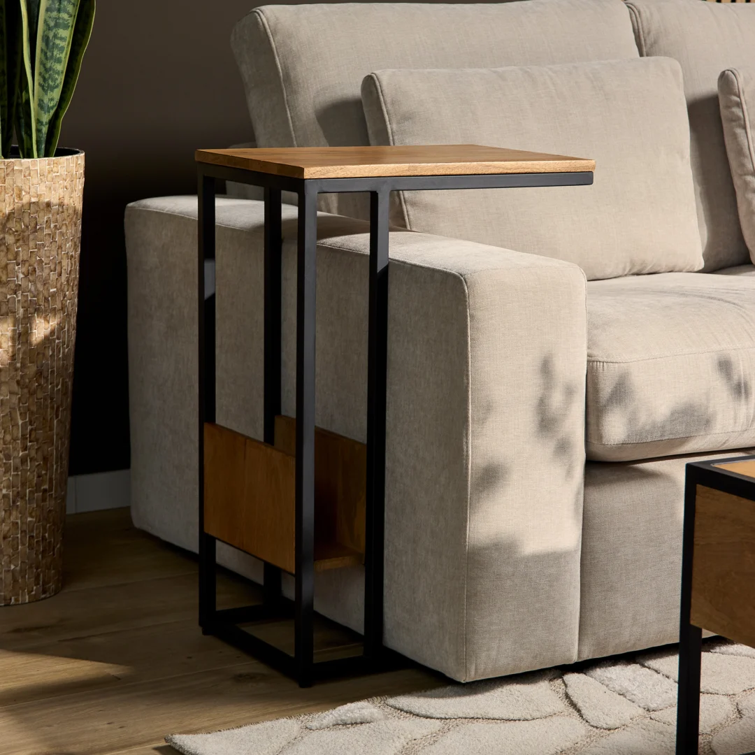 De Schuifregelaar Naturel Hoog, een moderne bijzettafel met een houten blad en een zwart metalen frame, staat naast een beige bank. Een plantenpot met textuur staat dichtbij en het zonlicht werpt zachte schaduwen op de bank en de tafel.