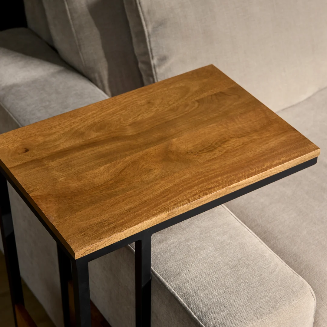 De Schuifregelaar Naturel Hoog, een compacte houten bijzettafel met een metalen frame, is geplaatst over de arm van een lichtgrijze stoffen bank.