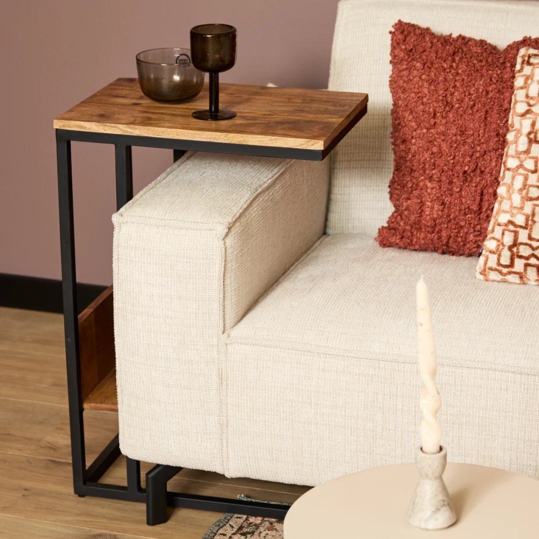 De Schuifregelaar Naturel Hoog, een modern bijzettafeltje van hout en metaal, past over een lichte bank met twee rood-oranje kussens met textuur, waarop een glas en een kopje staan. Een gedraaide kaars staat op een rond tafeltje op de voorgrond.