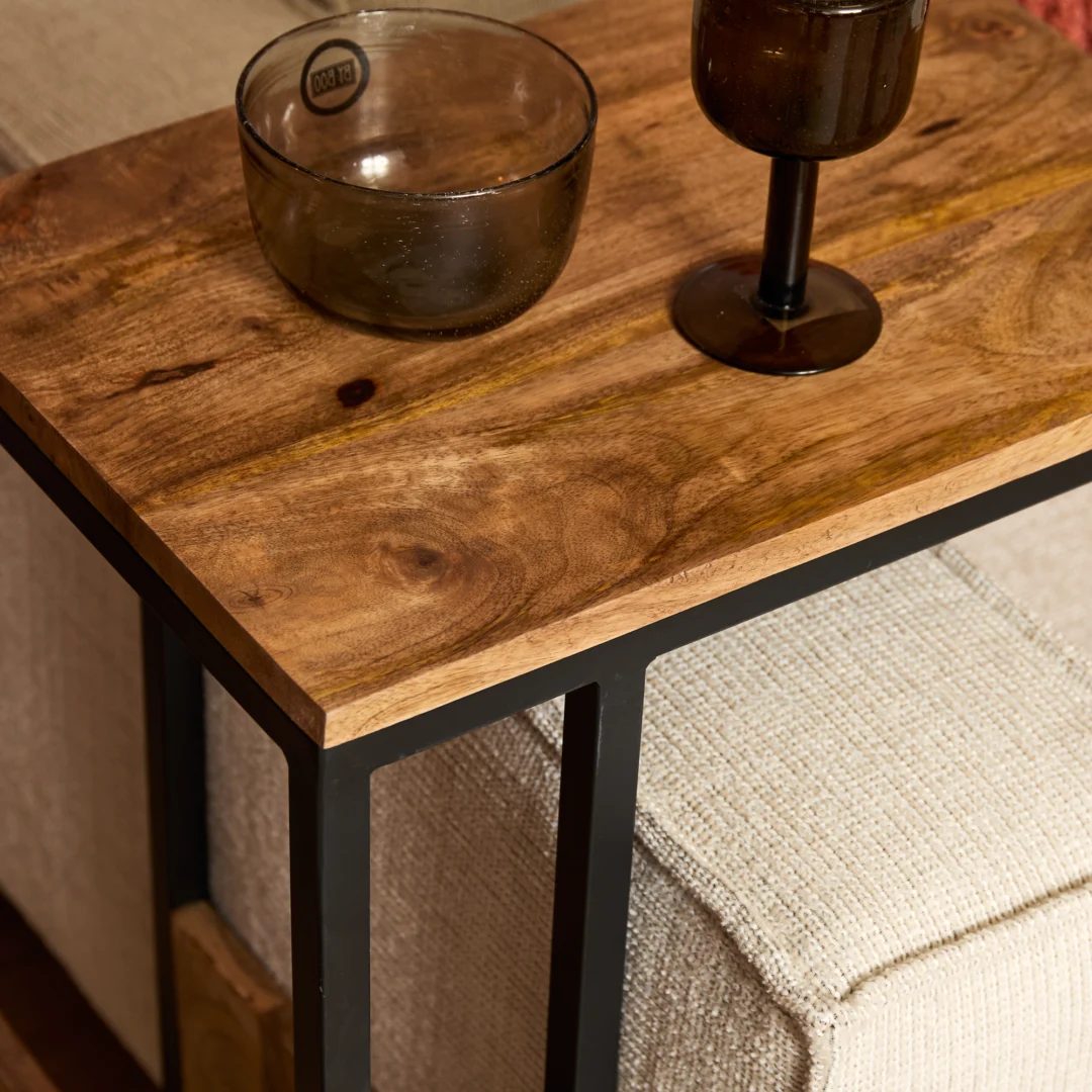 Een close-up van de Schuifregelaar Naturel Hoog, een houten bijzettafel met een zwart metalen frame, naast een lichte stoffen bank. Op het tafeltje staan een transparant glazen schaaltje en een donker glazen bokaal.