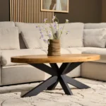 Een moderne woonkamer met een Salontafel 10 x 76 CM Ovaal, met zwarte metalen poten, een vaas met paarse bloemen erop, een lichtbeige sofa en neutrale decoratie op de achtergrond.