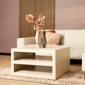 Een moderne woonkamer heeft een crèmekleurige bank met gestructureerde kussens, een Salontafel Zand 86 x 86 CM Vierkant salontafel en een vaas met roze bloemen. De achtergrond bestaat uit een houten lattenwand en een vloerkleed met patroon.