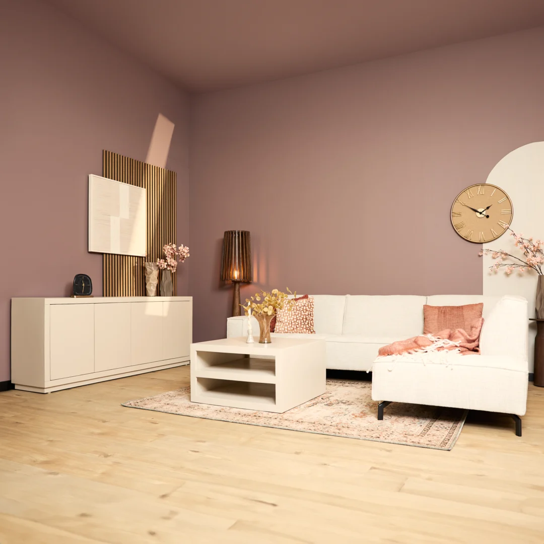 Een moderne woonkamer met lichthouten vloeren, mauve muren, een witte sectionele bank met roze kussens, een Salontafel Zand 86 x 86 CM Vierkant salontafel, dressoir, muurkunst, staande lamp, grote wandklok en decoratieve bloemen.