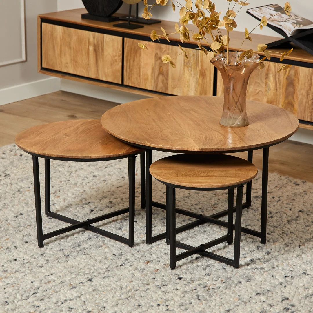 Houten salontafel set van drie, rond, met zwart metalen onderstel op een zacht kleed. Vaas met takken. Warme, natuurlijke belichting.