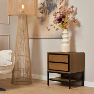 Houten nachtkastje met lade, staande lamp en vaas met bloemen. De nachtkast heeft een metalen frame. Zachte, natuurlijke kleuren en warme belichting.