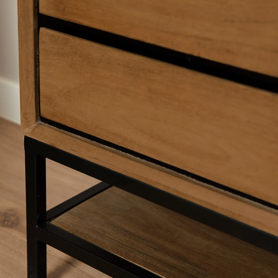 Detailopname van een houten nachtkastje met lade, zwart metalen frame en plank. Zachte belichting, warme houttinten.