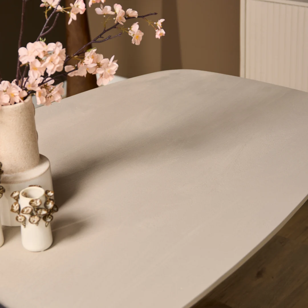 De Eettafel Madison Sand (160 cm, Deens Ovaal) toont minimalistische keramische vazen met roze bloesems. Neutraal gekleurde muren en een houten vloer maken de rustige, moderne omgeving compleet.