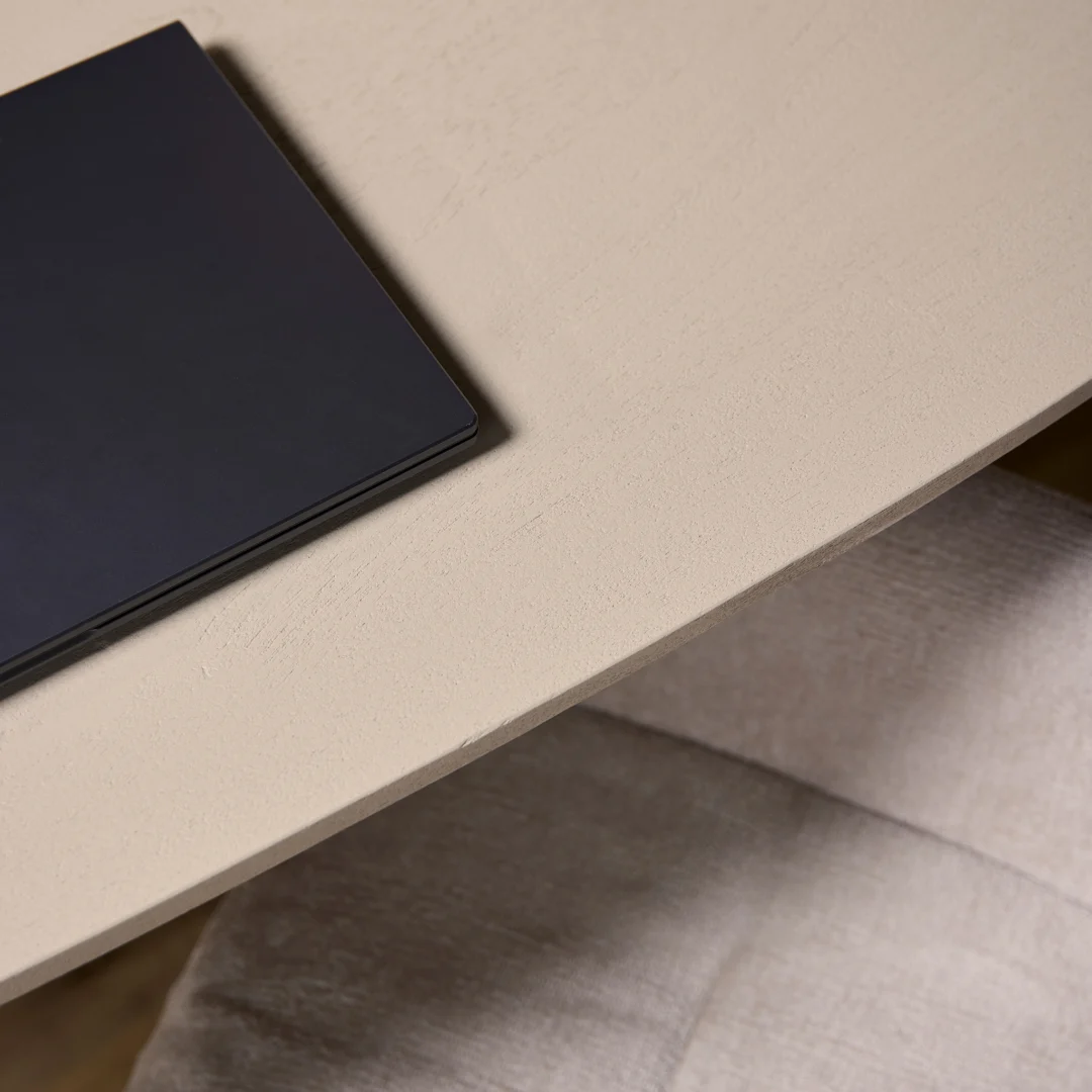 Een close-up van de Eettafel Madison Sand (240 cm, Deens Ovaal) beige tafelblad met een gesloten zwarte laptop in de linkerhoek en een lichtgekleurde stoel met kussens eronder.