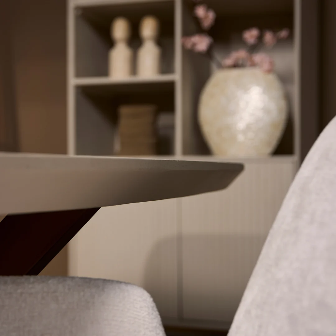 Een close-up van de Eettafel Madison Sand (240 cm, Deens Ovaal) met een moderne beige stoel, tegen een vage achtergrond met planken met decor, gestapelde boeken, twee beeldjes en een witte vaas met roze bloemen.