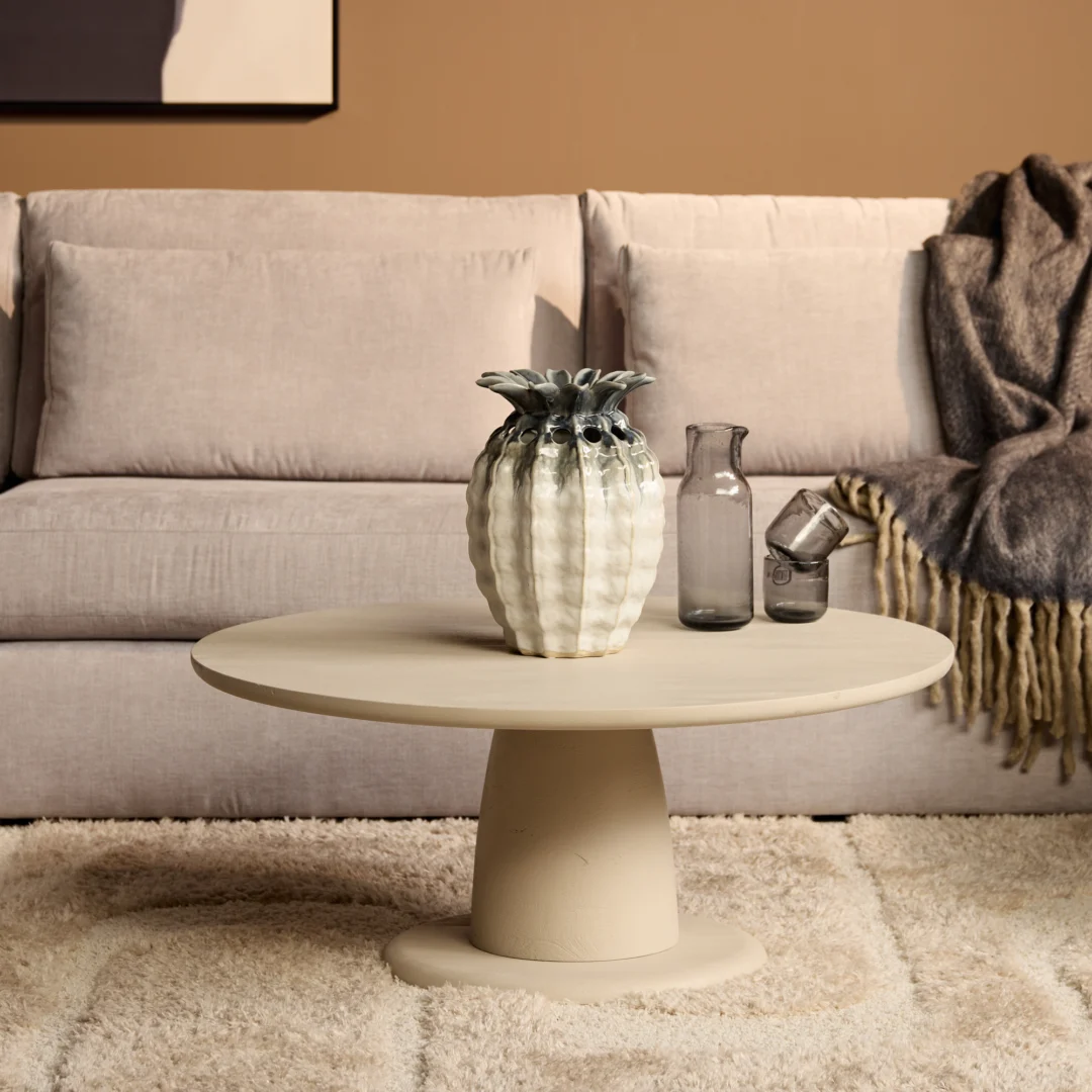 Beige ronde salontafel met vaas en glazen. Zachte belichting, beige kleurenpalet. Sofa en kleed op de achtergrond. Minimalistisch design.