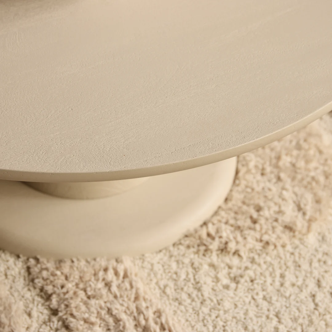 Detailopname van een ronde, crèmekleurige salontafel op een zacht tapijt. Het tafelblad is glad en het licht is zacht en diffuus.