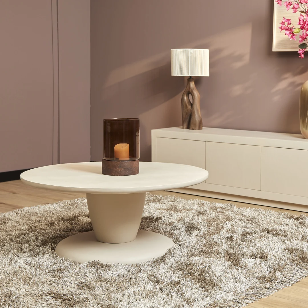 Beige ronde salontafel met kaars in een glazen houder. Zachte belichting, beige vloerkleed en kast. Warme, neutrale kleuren.