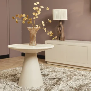In een moderne woonkamer staat de Salontafel Roest 67 x 64 CM als ronde bijzettafel met een glazen vaas met gouden takken. Daarachter staan een wit dressoir en een sculpturale lamp tegen een mauve muur op een beige vloerkleed met textuur.