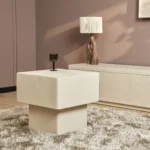 In een moderne woonkamer staat de Salontafel 57 x 52 CM Vierkant, een vierkante crèmekleurige tafel op een ruig tapijt, met een donker glas bovenop, een wit dressoir, een sculpturale lamp en mauve muren.