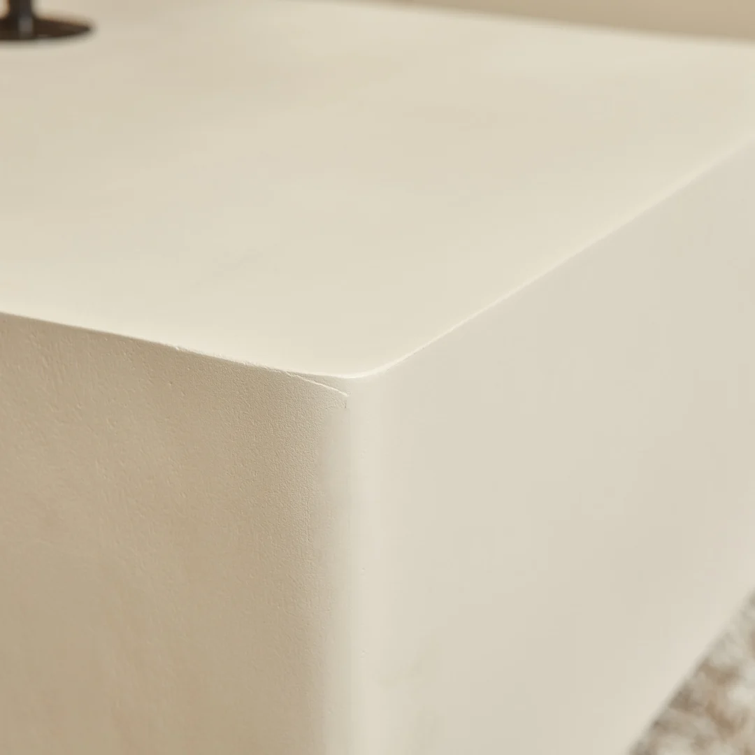 Close-up van de Salontafel 57 x 52 CM Vierkant, met een glad, gebroken wit vierkant blok met afgeronde hoeken en een licht gestructureerd oppervlak; een deel van een ander meubelstuk verschijnt op de achtergrond.