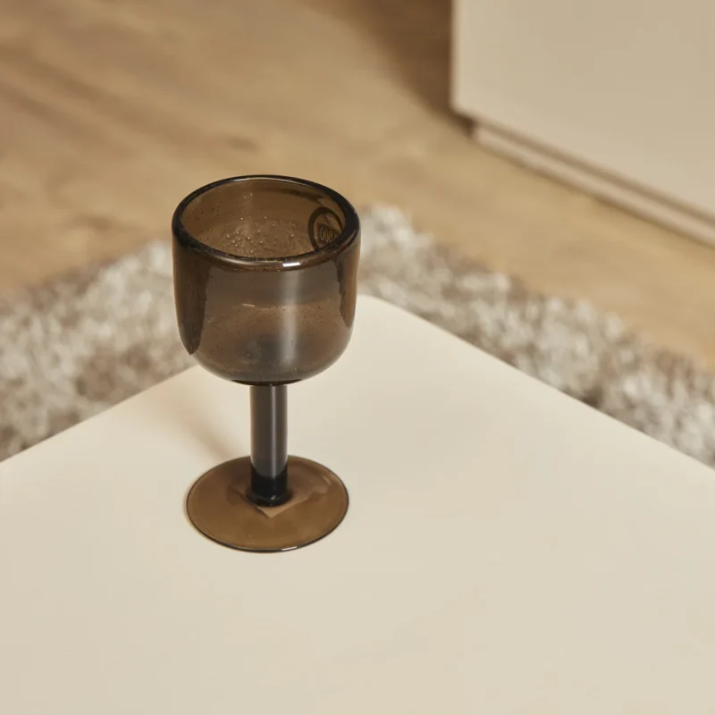 Een bruingetinte glazen bokaal staat op een witte Salontafel 57 x 52 CM Vierkant, met een vaag beige en grijs tapijt en houten vloer op de achtergrond.