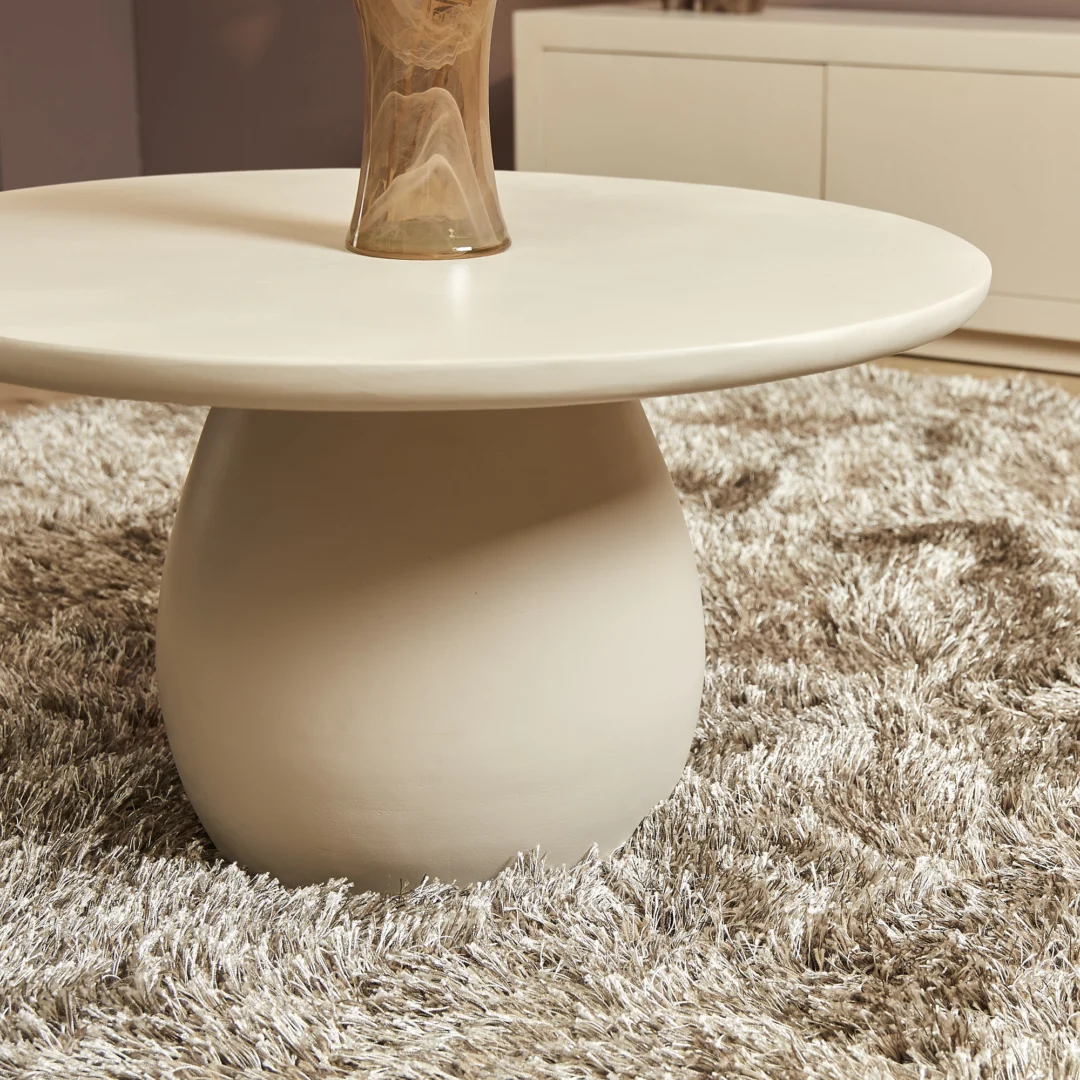De Salontafel 70 CM Vierkant, een moderne witte vierkante salontafel met een glad onderstel, staat op een beige shagtapijt. Een bruine glazen vaas is gedeeltelijk zichtbaar op het tafelblad, met een witte kast op de achtergrond.