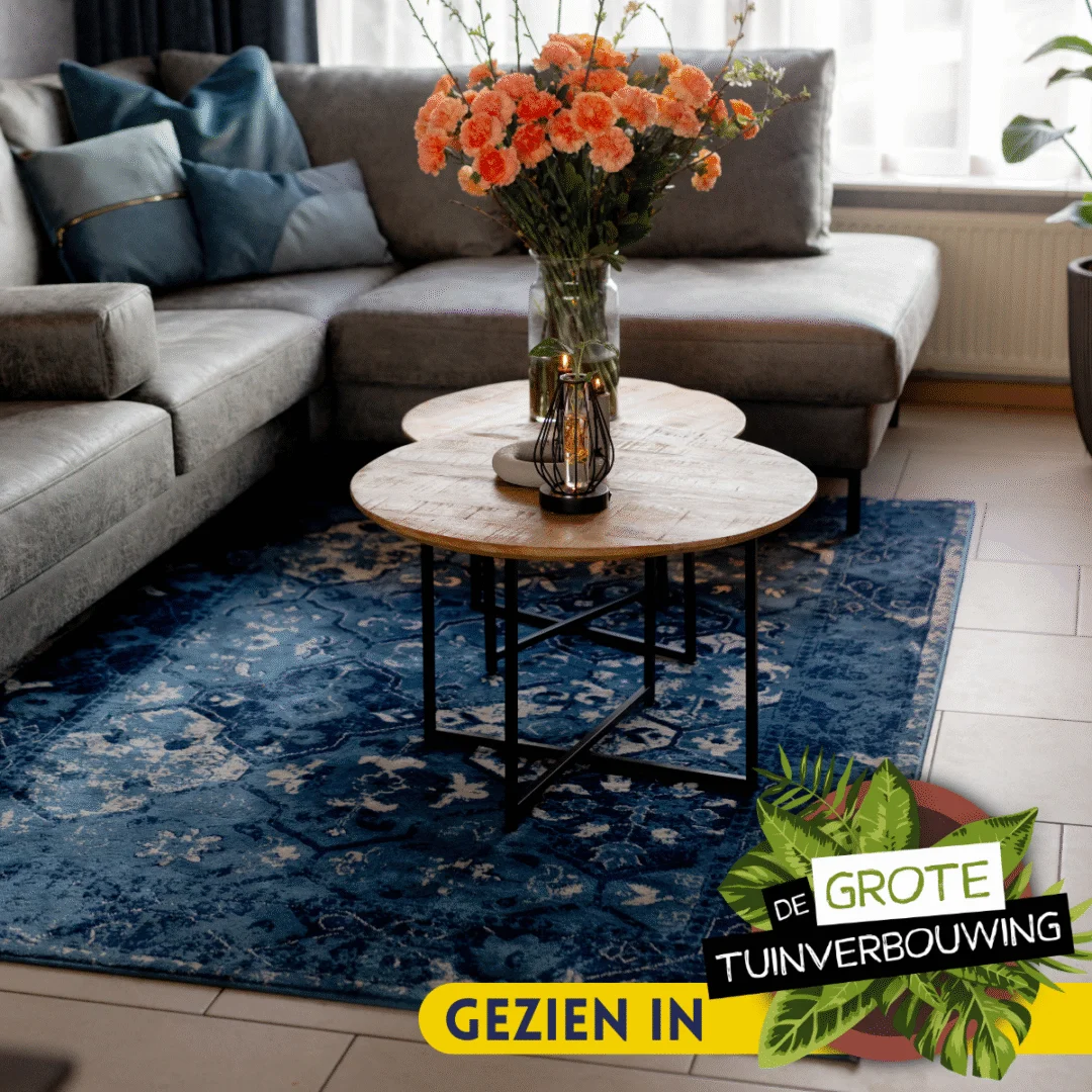 Een moderne woonkamer met een grijze sectionele bank, een blauw gedessineerd vloerkleed en een Salontafel 60 CM met daarop oranje bloemen in een vaas. Op een decoratieve banner in de hoek staat GEZIEN IN De Grote Tuinverbouwing.