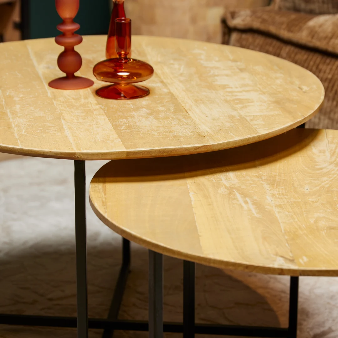 Twee ronde Salontafel 60 CM met zwart metalen poten zijn gedeeltelijk in elkaar geschoven. Twee amberkleurige glazen vazen in verschillende vormen staan op de grotere tafel, met een bruine stoel zichtbaar op de achtergrond.