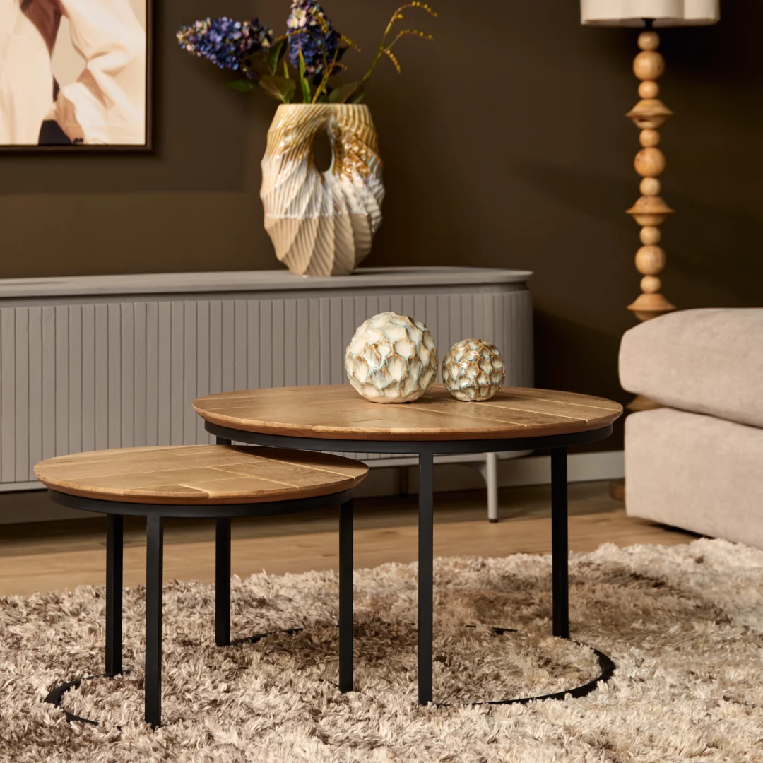 Twee ronde salontafels van eikenhout met zwarte poten op een zacht kleed. Decoratieve vazen en een lamp op de achtergrond. Warm en sfeervol.