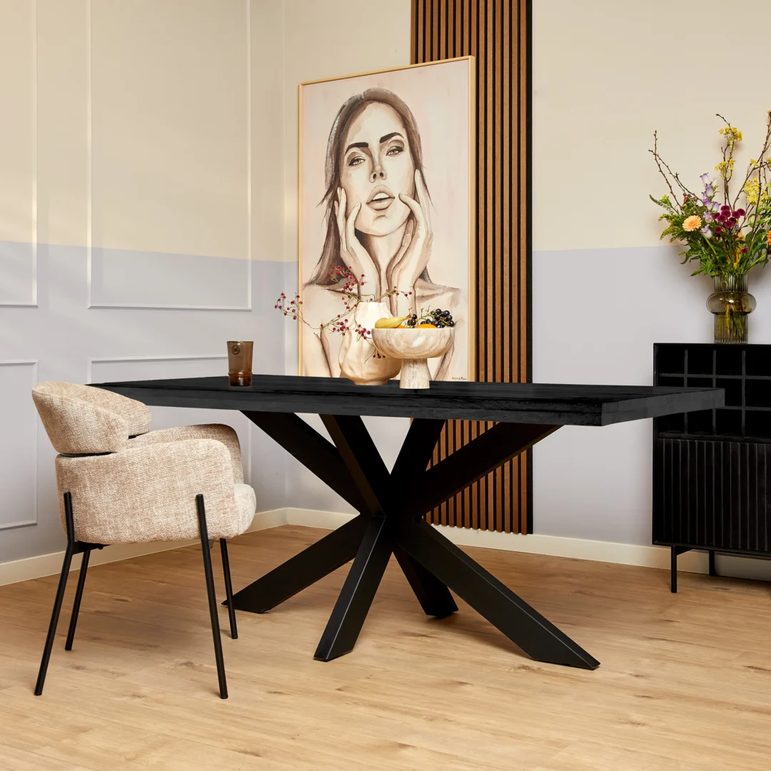 Een moderne eetkamer met de Eettafel Denver Zwart (200 cm), beige stoelen met kussens, een fruitschaal en een drankje. Achteraan vullen een groot vrouwenportret en een bloemenvaas op een zwart dressoir de lichte houten vloer en neutrale muren aan.