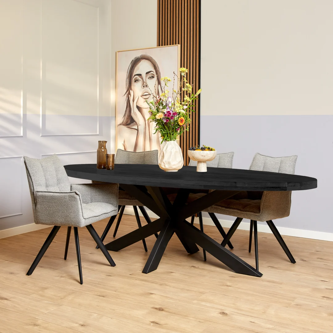 In een moderne eetkamer staat de Eettafel Denver Zwart Ovaal 210 cm met vier grijs gestoffeerde stoelen, een vaas met kleurrijke bloemen, decoratieve vazen en een schaal. Een groot vrouwenportret leunt tegen een neutrale muur op een houten vloer.