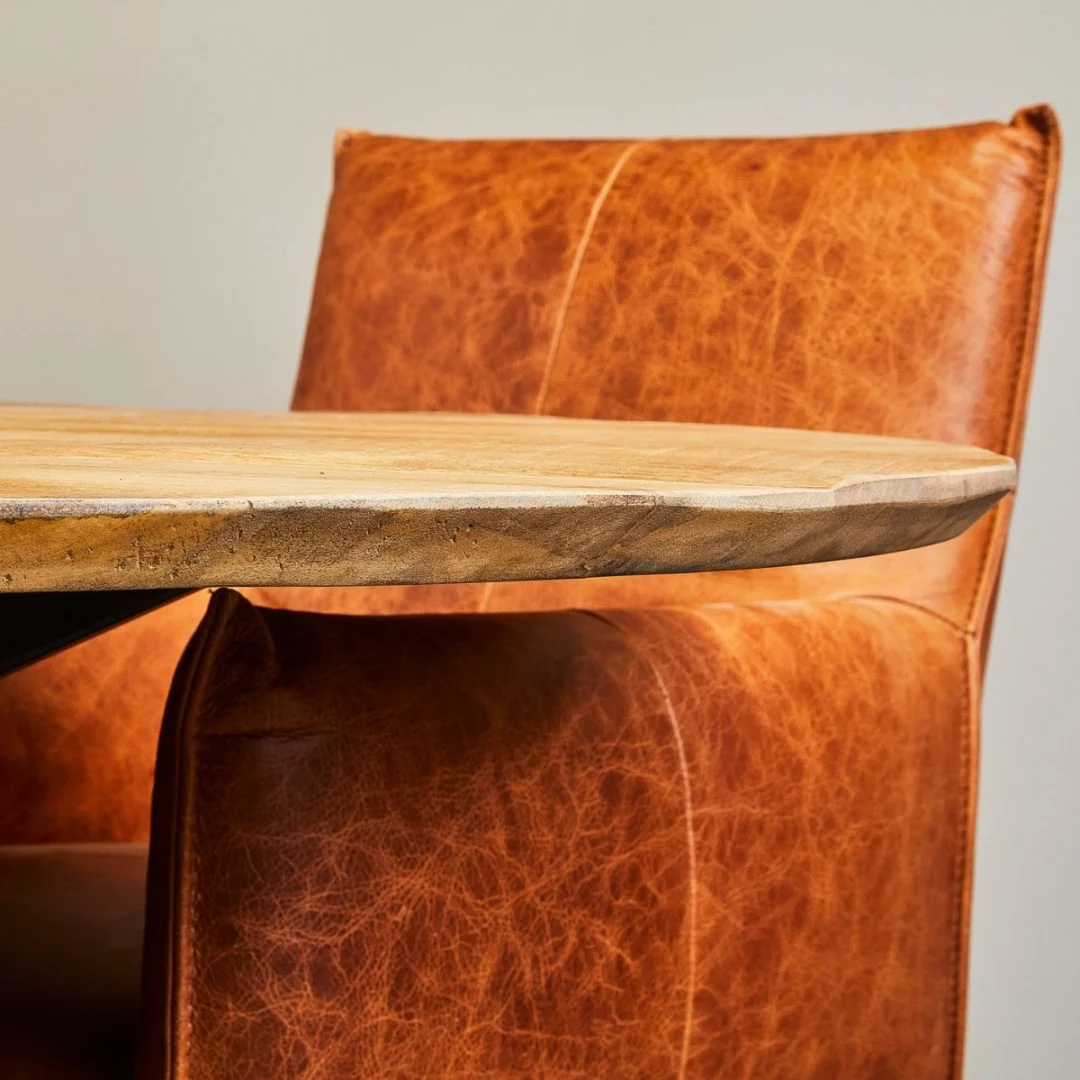 Een close-up van de Eettafel Arlington | Rond | 120 cm met zijn ronde houten rand naast een beklede bruinleren stoel met zichtbare stiksels, tegen een effen, lichtgekleurde muur.