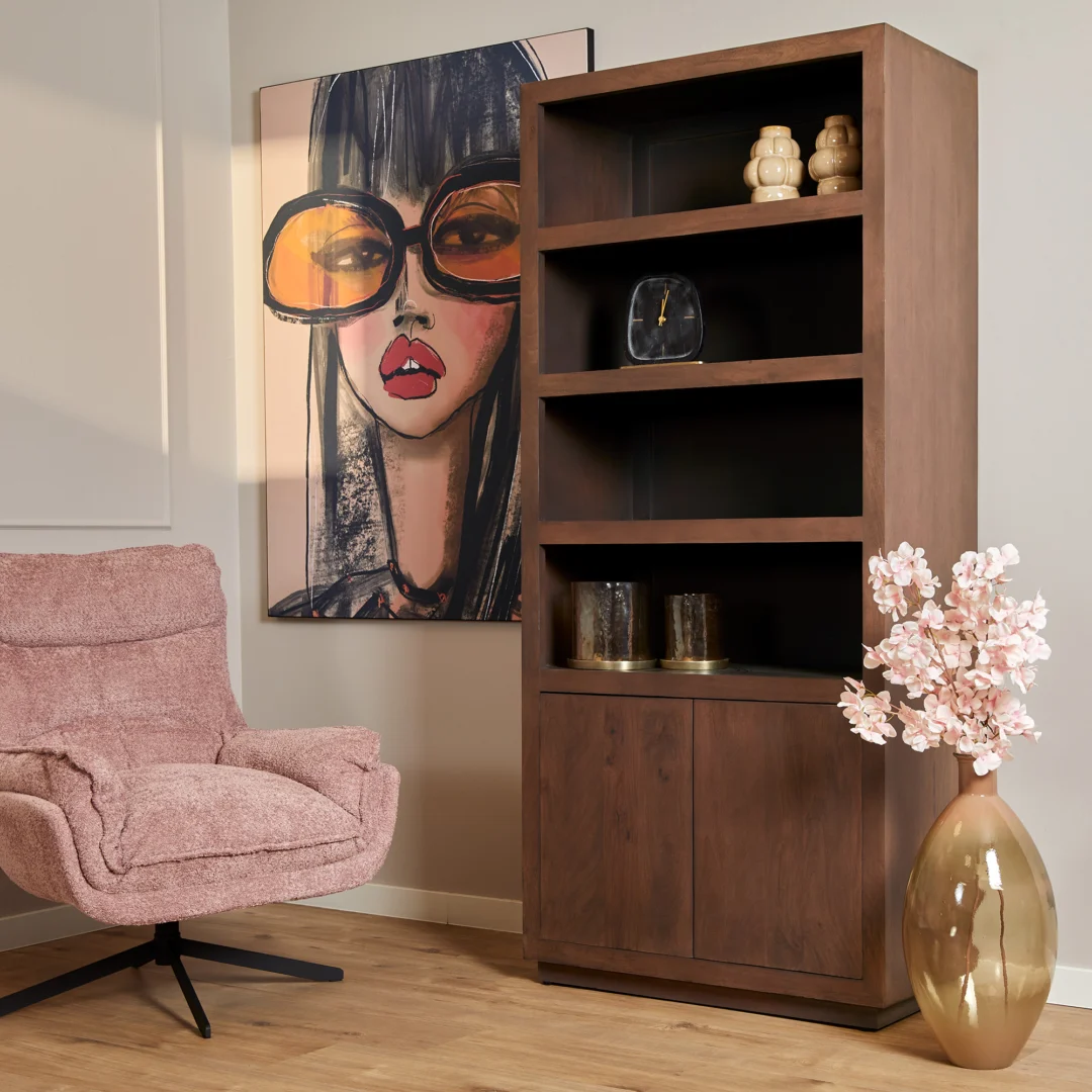 Een moderne woonkamerhoek met een pluche roze stoel, Salontafel Walnoot Compact, een houten boekenplank met decor, een groot schilderij van een vrouw met een te grote bril en een hoge gouden vaas met roze bloemen op een houten vloer.