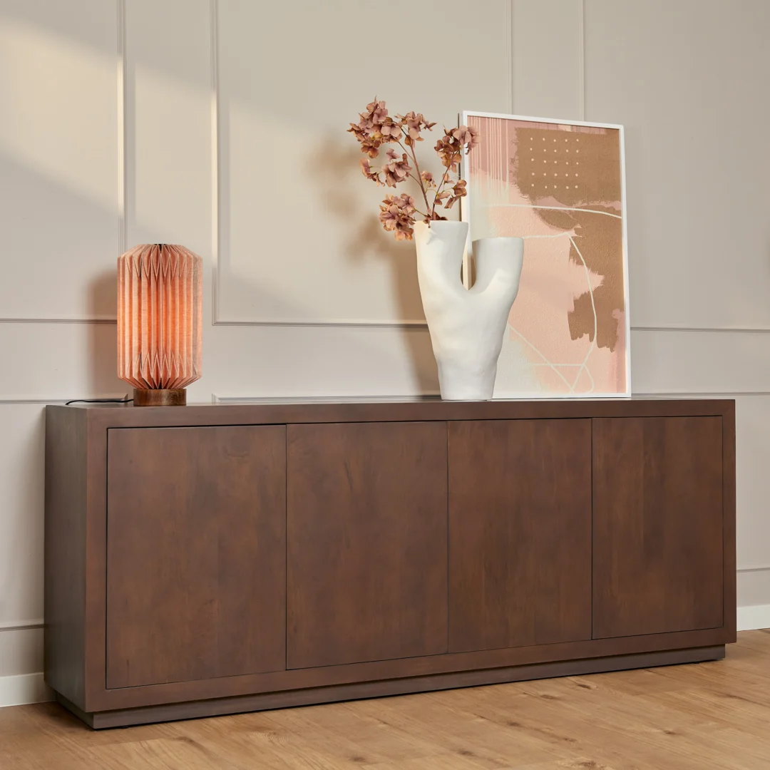 De Salontafel Walnoot Compact, een moderne houten tafel, staat tegen een muur met beige panelen met daarop een roze lamp, een sculpturale witte vaas met gedroogde bloemen en een abstracte print in bruin- en bloostinten.