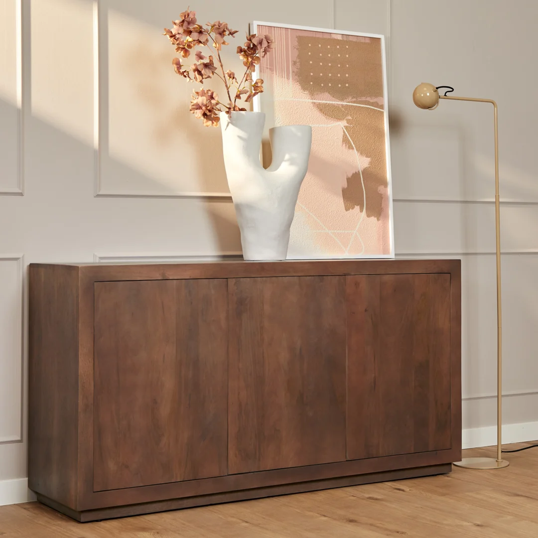 De Salontafel Walnoot Compact staat tegen een beige wand met panelen, met daarop een sculpturale witte vaas met roze bladeren en een ingelijste abstracte print. Een hoge gouden vloerlamp met een ronde lamp staat naast de tafel.