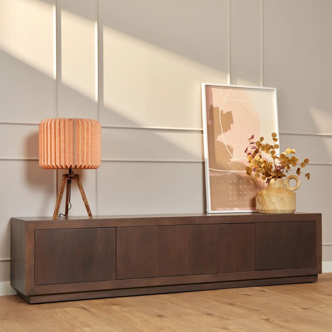 De Salontafel Walnoot Compact, een modern houten dressoir met vier laden, staat tegen een muur met beige panelen. Bovenop staan een roze tafellamp, ingelijste abstracte kunst en een decoratieve vaas gevuld met gedroogd geel gebladerte.