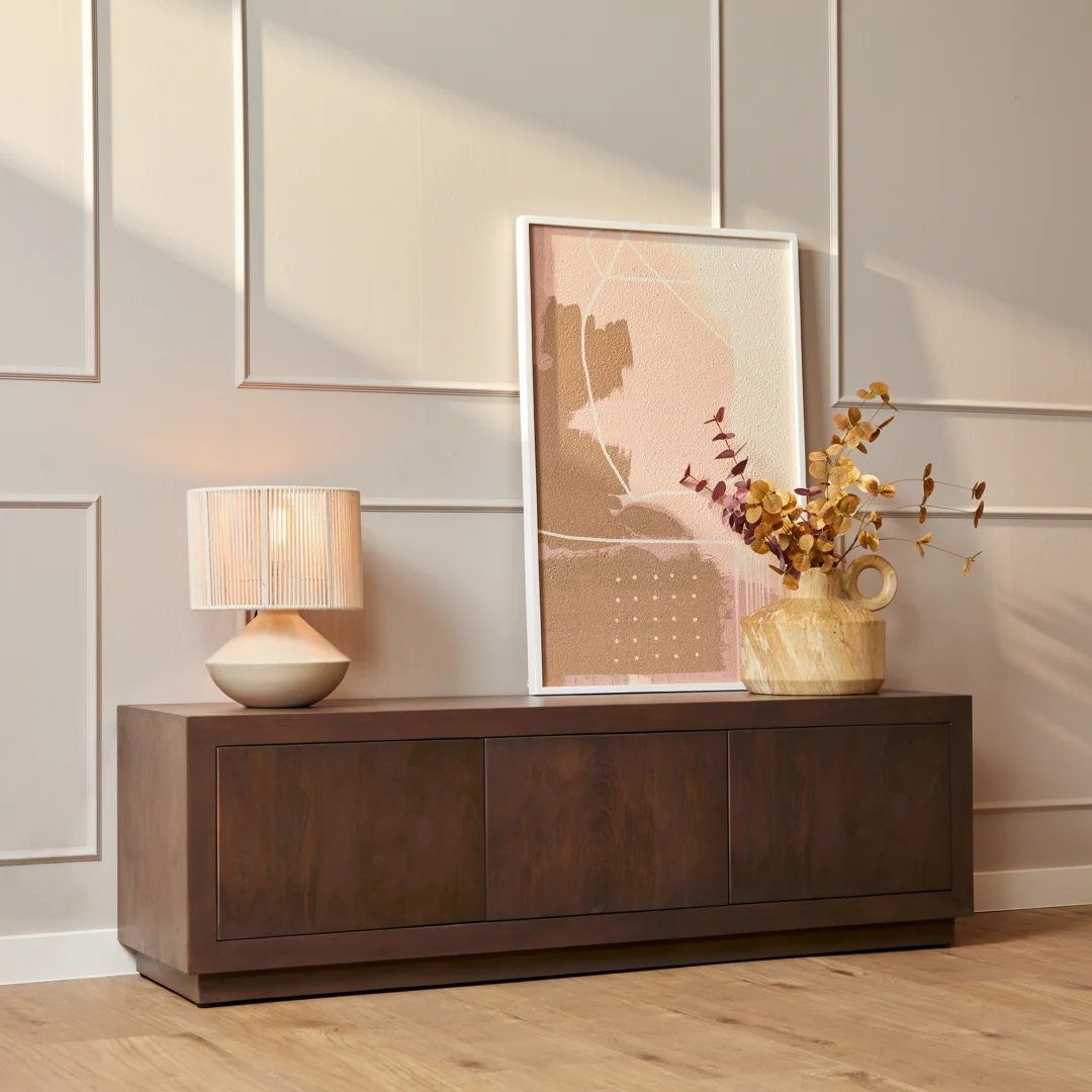 De Salontafel Walnoot Compact staat tegen een beige wand met panelen, met daarboven een tafellamp met geplooide kap, minimalistische abstracte kunst en een vaas met gedroogde gouden takken. Een licht houten vloer maakt de moderne look compleet.