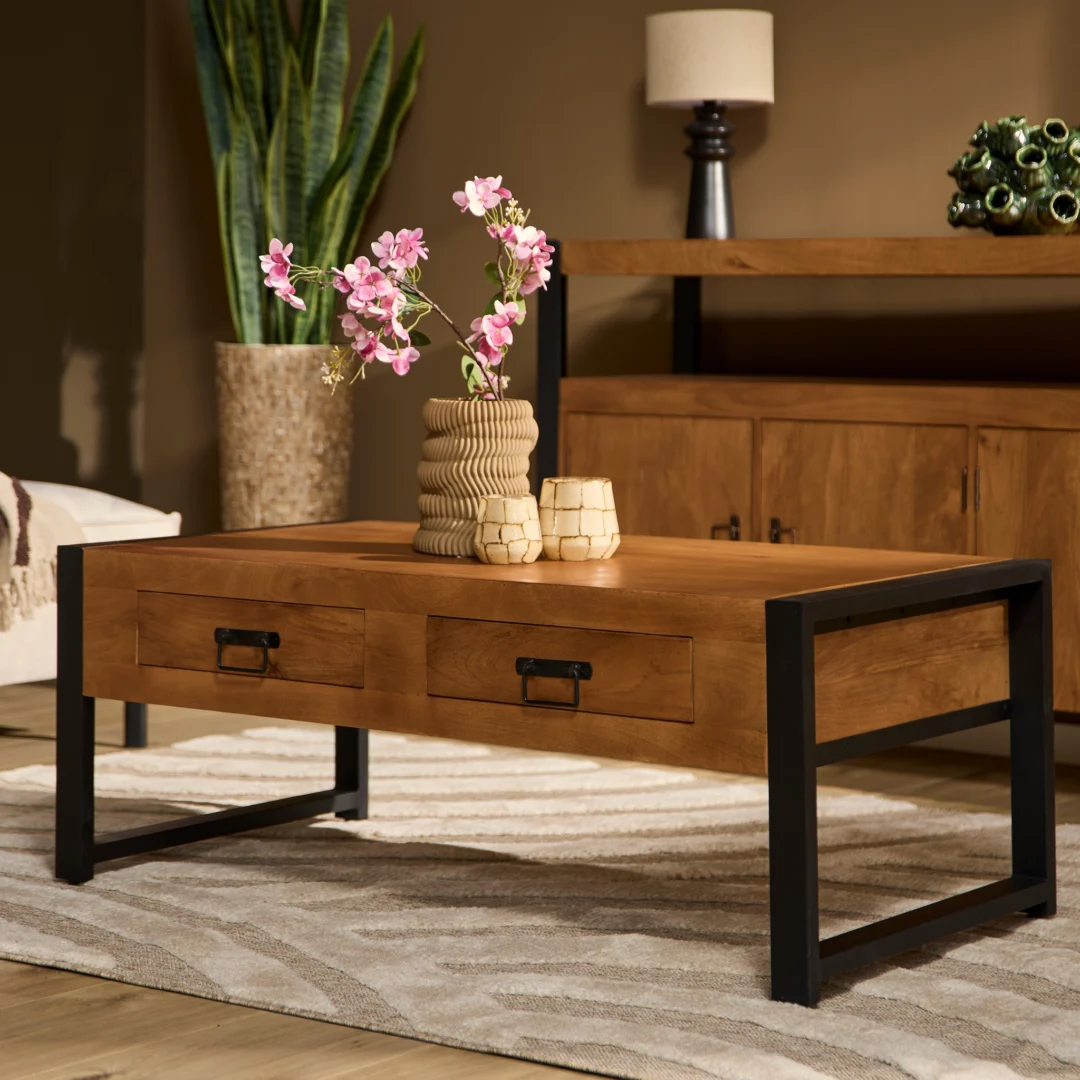 De Salontafel 120 x 60 CM Design heeft een moderne houten afwerking, zwarte metalen poten en twee lades. Bovenop staan structuurvazen met roze bloemen. Op de achtergrond staan een houten dressoir, een hoge groene plant en een lamp.