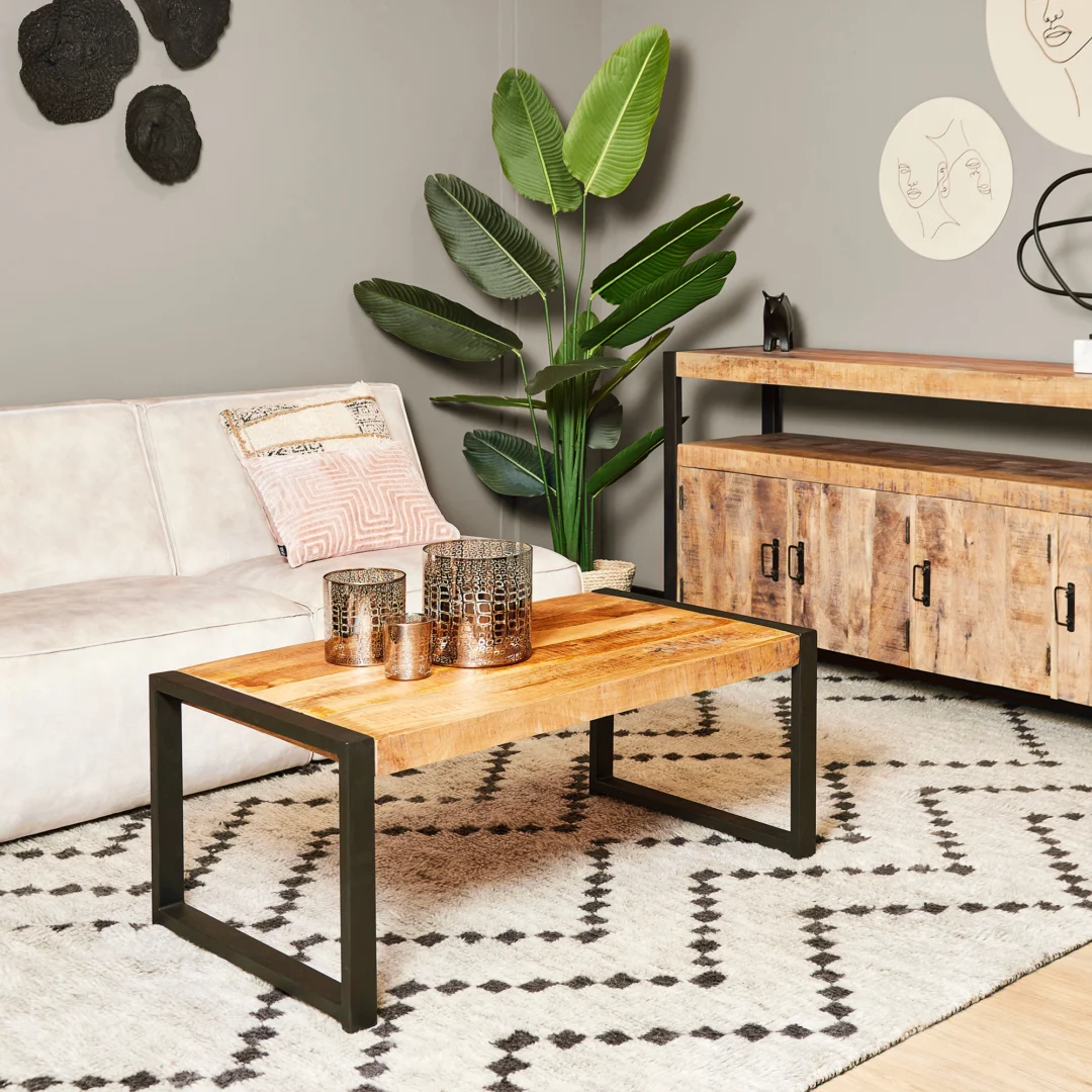 Een moderne woonkamer met een beige bank, Salontafel 60 x 45 CM (houten salontafel met metalen poten), vloerkleed met patroon, dressoir, grote groene plant, decoratieve kaarsen en minimalistische muurkunst op een lichtgrijze muur.