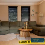 Een gezellige woonkamer met een donkergroen bankstel, de Salontafel 70 x 70 CM Rond met kaarsen, ronde vloerkleden, muurkunst en twee ramen met zwarte jaloezieën. Tekst: "Gezien in De Grote Huisverbouwing.