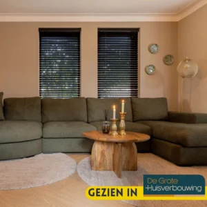 Een gezellige woonkamer met een donkergroen bankstel, de Salontafel 70 x 70 CM Rond met kaarsen, ronde vloerkleden, muurkunst en twee ramen met zwarte jaloezieën. Tekst: "Gezien in De Grote Huisverbouwing.