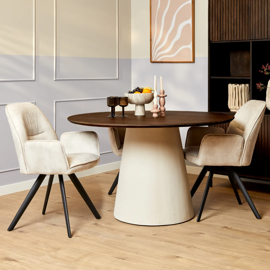 Elegante Madison eettafel met ronde houten blad en crèmekleurige voet. Beige stoelen met zwarte poten op een houten vloer. Sfeervolle belichting.