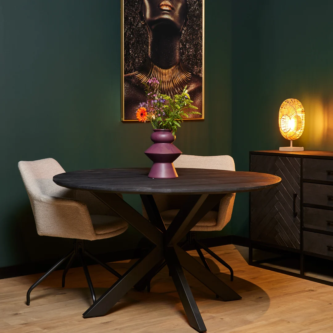 Een moderne eethoek met de Eettafel New York ronde (110 cm) zwarte tafel, twee beige stoelen, een paarse vaas met bloemen, een decoratieve lamp op een zwarte kast en een ingelijst vrouwenportret op een donkergroene muur.