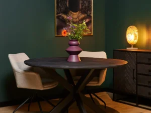 Een moderne eethoek met de Eettafel New York Rond 130 cm in zwart, twee beige stoelen, een paarse bloemenvaas, een dressoir met een brandende lamp en vrouwenkunstwerk met goudaccenten op een donkergroene muur.