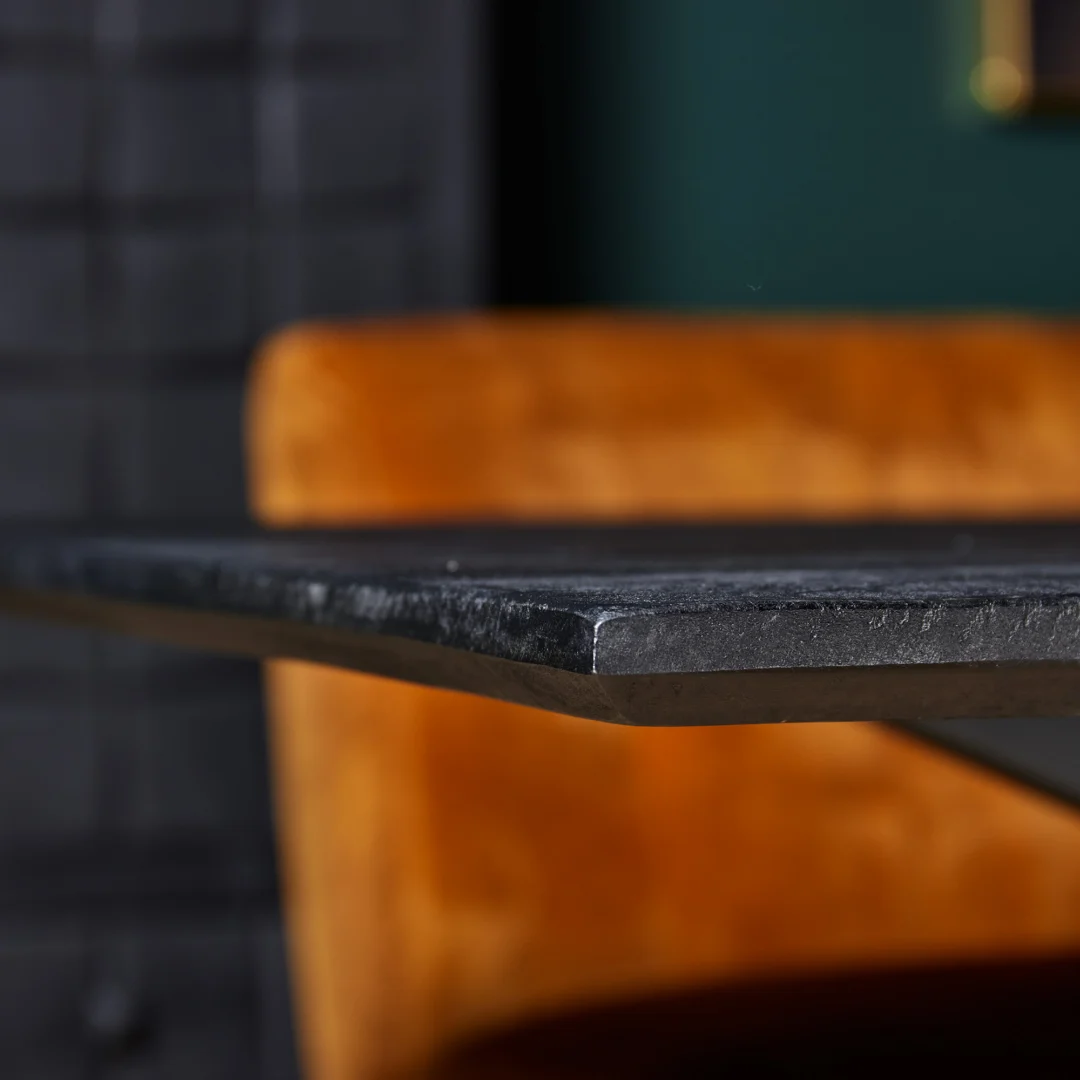 Close-up van de Eettafel New York | Rechthoek | 200 cm met een donker gestructureerd oppervlak; een vage oranje stoel en groene muur op de achtergrond creëren een moderne, stijlvolle interieursfeer.