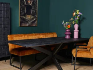 Een moderne eetkamer met de Eettafel New York Rechthoek 180 cm zwarte tafel, een oranje fluwelen bank en stoel, twee roze bloemenvazen, donkergroene muren, een houten vloer en een groot vrouwenportret met gouden accenten aan de muur.