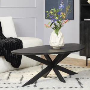 De Salontafel 130 x 70 CM Rond Compact, een zwarte ovale salontafel met een modern geometrisch onderstel, staat op een wit gedessineerd vloerkleed. Op de tafel staat een witte vaas met kleurrijke bloemen; een witte bank en zwarte deken verschijnen op de achtergrond.