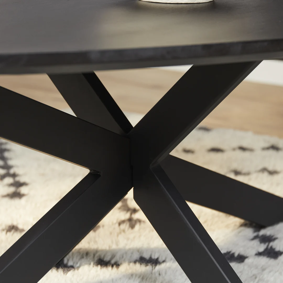 Close-up van het zwart metalen onderstel met kruisende poten van de Salontafel 130 x 70 CM Rond Compact op een wit en zwart gedessineerd tapijt, met een deel van het donkere tafelblad en een klein object erboven.